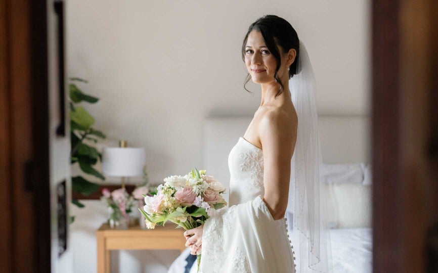 A bride in an elegant strapless gown and veil holds a bouquet of pastel flowers while smiling softly inside a bright, stylish room at The Wilbur Mansion.