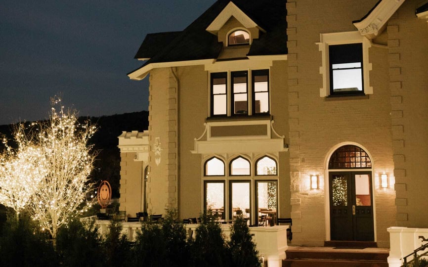 The Wilbur Mansion illuminated at night, with glowing string lights on trees and warm light shining through its arched windows and doorway