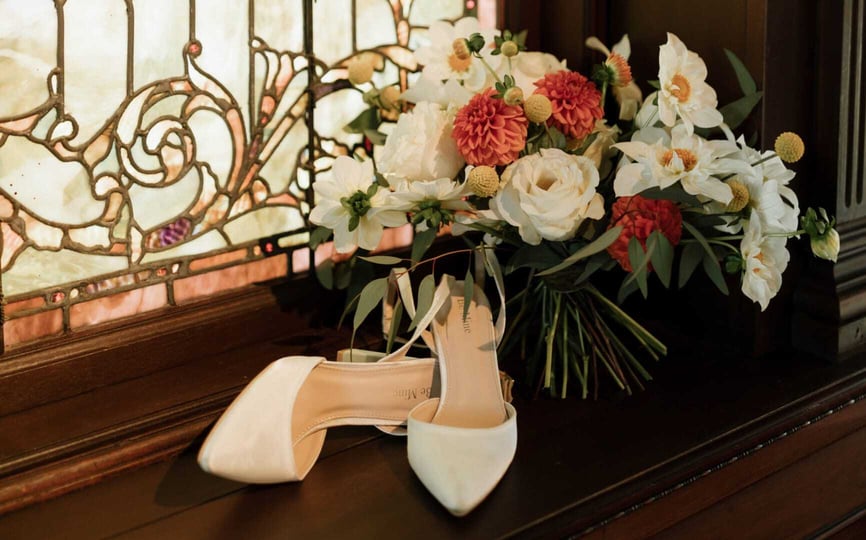 A bridal bouquet of white and coral flowers rests beside ivory heels on a wooden windowsill with a stained-glass backdrop at The Wilbur Mansion.
