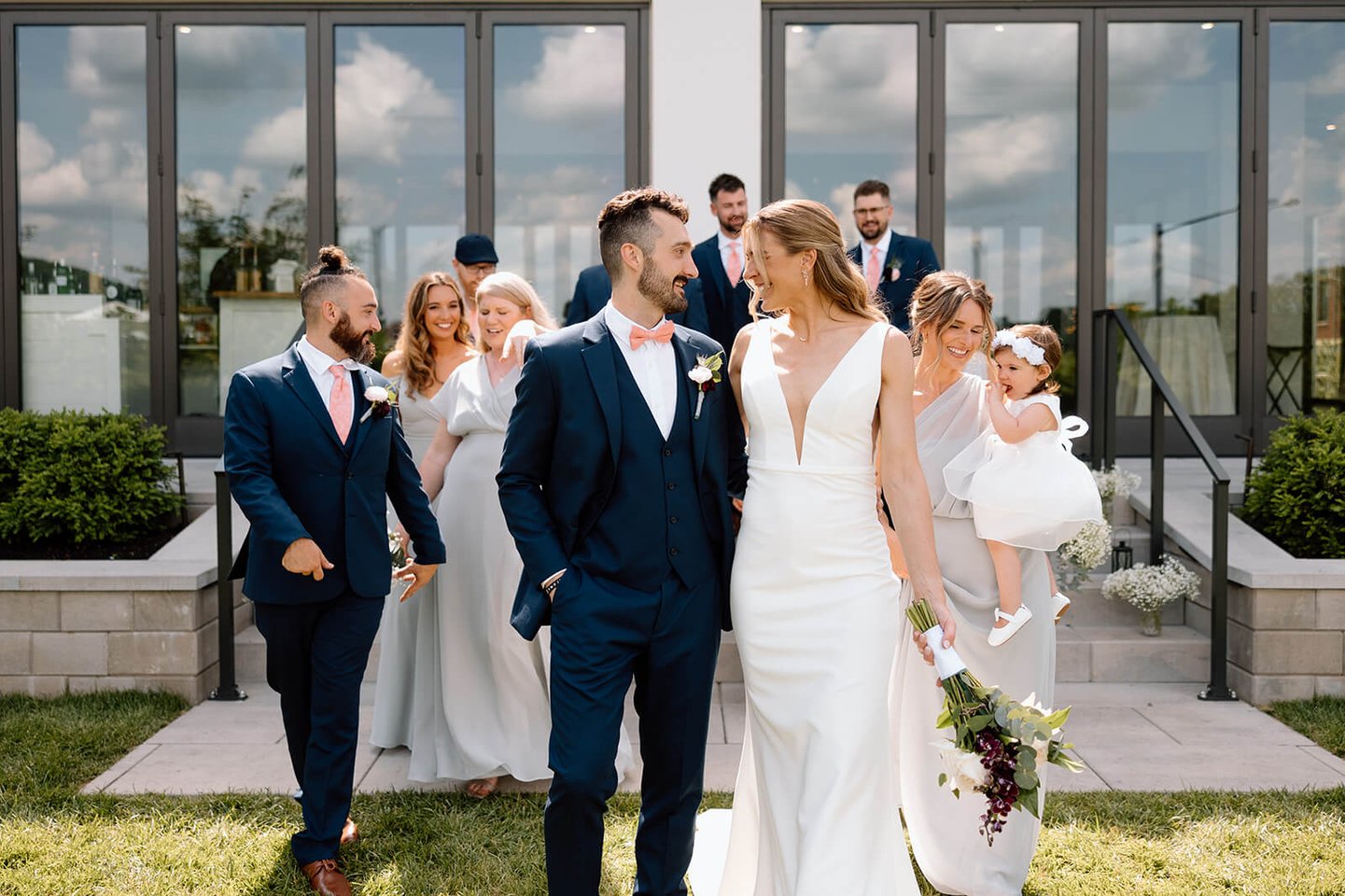 A bride and groom smile at each other while walking with their wedding party outside The Wilbur Mansion, surrounded by bridesmaids, groomsmen, and a flower girl in white