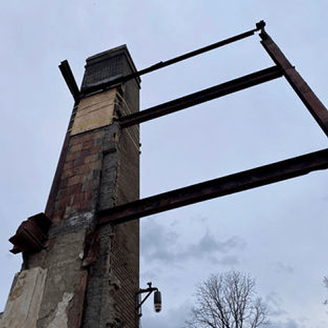 A tall brick chimney supported by steel beams during restoration work at The Wilbur Mansion site, set against a cloudy sky.