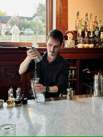 A bartender prepares a cocktail at The Wilbur Mansion bar, pouring a drink from a shaker into a glass on a marble counter surrounded by bottles and bar tools.