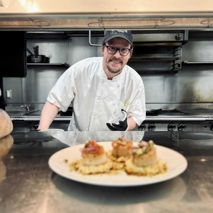 A chef in a white uniform and black cap smiles in a professional kitchen, presenting a plated gourmet dish on the counter.