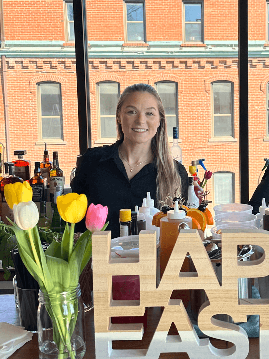A smiling bartender stands behind a bar setup with colorful tulips, drink mixers, and bottles, with a wooden “HAPPY EASTER” sign in the foreground and brick building windows in the background.